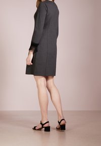 Grå långärmad klänning med figursydd form, med svarta spetsdetaljer vid ärmarna. Modellen har på sig svarta klacksandaler med blockklack.