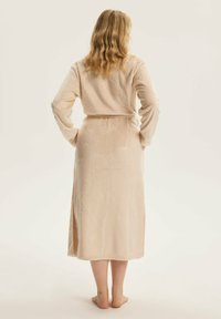 Długi, jasnobeżowy szlafrok o teksturowanym wzorze przypominającym kształty wachlarzy, z długimi rękawami i wiązanym w pasie. Miękki materiał sięga do połowy łydki.