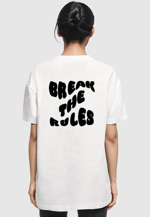 Hvid oversized t-shirt med fed, sort tekst der står "OVERTRÆD REGULERNE" i et legende, bølget skrifttype på ryggen.