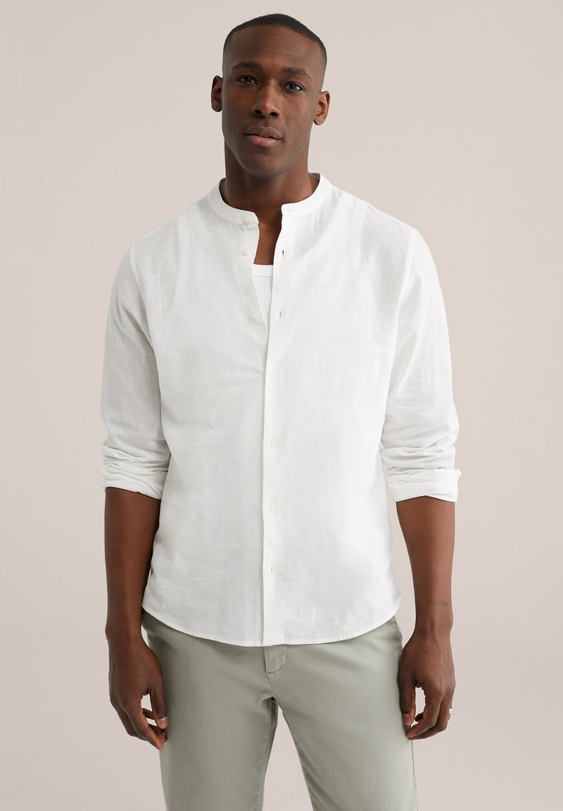 Muž stojící před jednobarevným pozadím, oblečený v bílé košili s knoflíky a dlouhými rukávy vyhrnutými až nad lokty a světle šedých kalhotách.