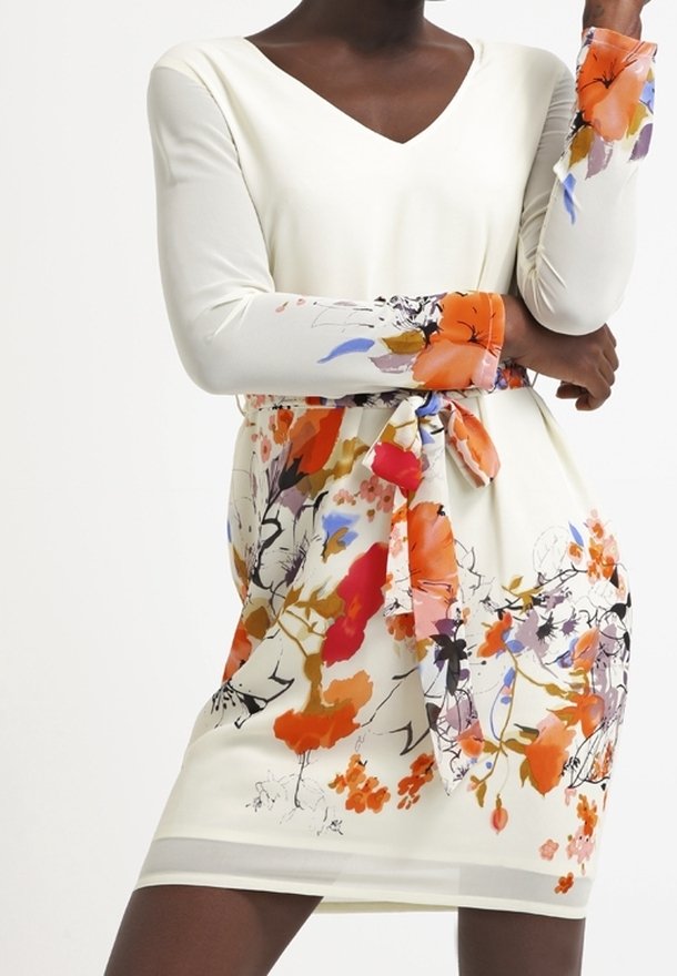Femme portant une robe blanche à manches longues avec des motifs floraux colorés orange, rouge et violet, et une ceinture assortie en tissu nouée à la taille.
