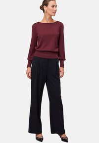 Top lungo bordeaux con scollo a barca, abbinato a pantaloni neri a gamba larga. Il look è completato da scarpe con tacco alto nere.