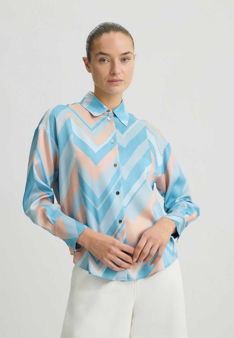 Cămașă colorată cu butoni, având un model în zig-zag în nuanțe de albastru, roz deschis și alb. Fabricată dintr-un material fin, cu mâneci lungi și guler.