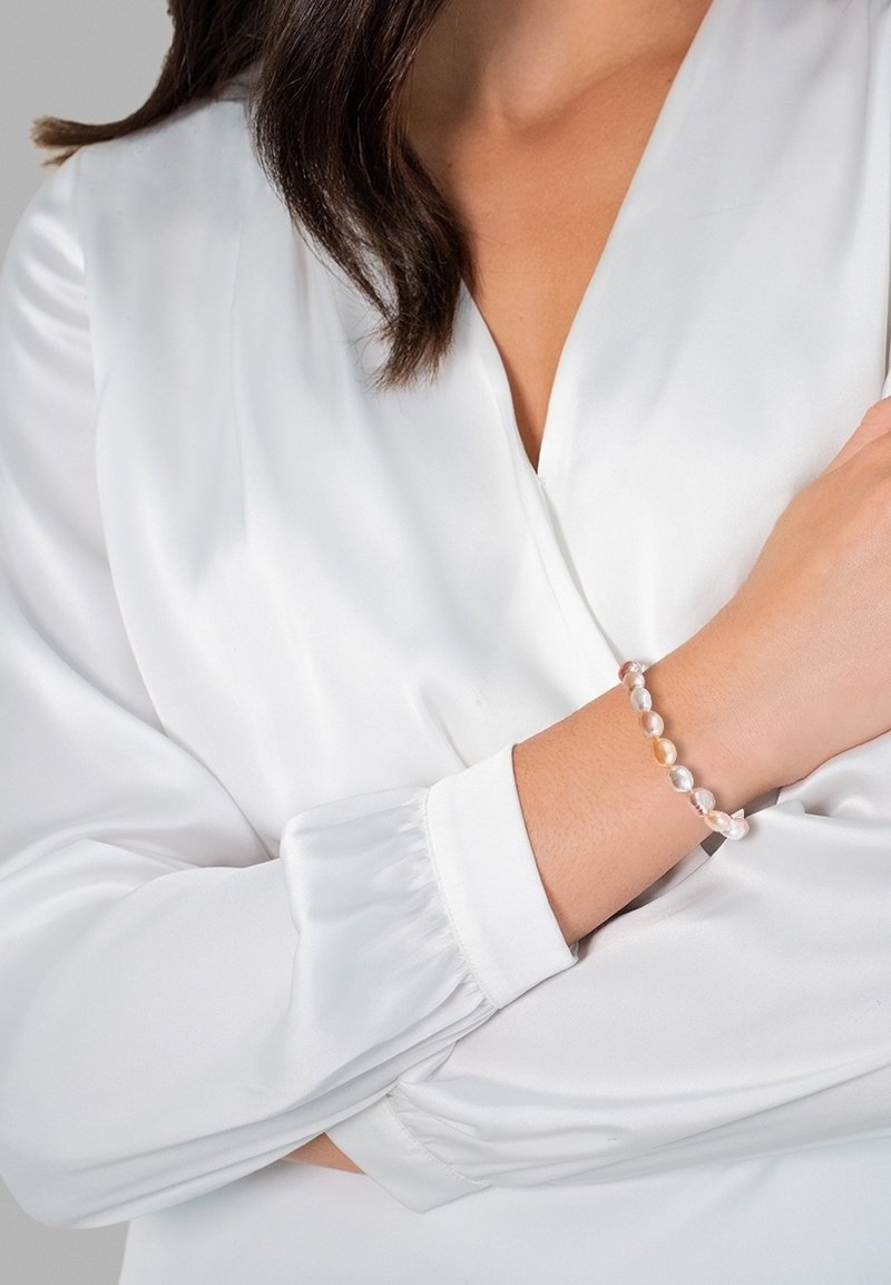 Pulsera de perlas con tamaños y tonos variados de perlas en rosa claro, crema y gris. Llevada en una muñeca, sobre una blusa de seda blanca.