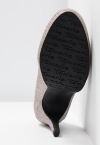 Zapato de tacón stiletto de color oro rosa brillante con punta afilada y suela negra texturizada que presenta el nombre de la marca en relieve.