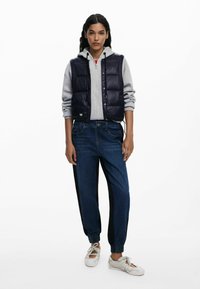 Chaleco acolchado navy sobre una sudadera gris, combinado con jeans jogger azules que tienen rayas laterales negras, y zapatillas blancas de cordones.