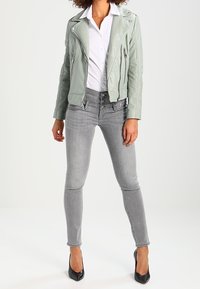 Chaqueta de motociclista de cuero verde claro, camisa blanca debajo, combinada con jeans ajustados grises y tacones altos negros. Cremalleras notables en la chaqueta.