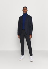 Marineblazer über einem blauen Rollkragenpullover, kombiniert mit dunklen Hosen und weißen Turnschuhen. Slim-Fit-Design, glattes Material und minimalistische Details.