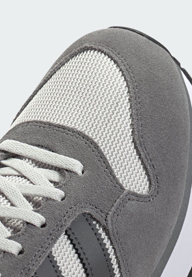 Grå atletisk sko med texturerad grå mockauppdel och ett meshnätparti, kontrasterande vita skosnören och mörkgrå ränder längs sidan.