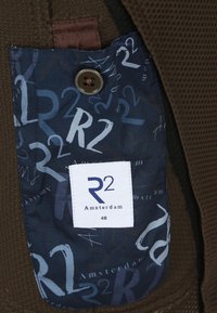 Braun strukturiertes Gewebe mit einer blauen gemusterten Tasche, die ein weißes "R2"-Logo und ein "Amsterdam 48"-Label zeigt, das mit einem Knopf gesichert ist.