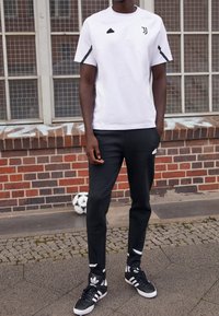 Biała koszulka sportowa z czarnymi akcentami i logiem, połączona z czarnymi zwężanymi spodniami oraz czarnymi trampkami Adidas z białymi paskami.