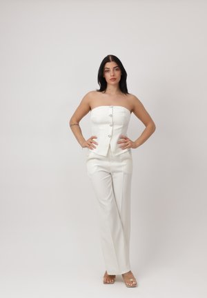 Joven mujer de cabello oscuro posa contra un fondo blanco, vistiendo un top blanco sin tirantes y pantalones blancos de cintura alta, con sandalias de tacón transparente.