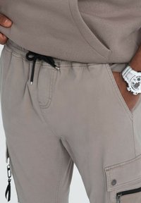 Nahaufnahme einer Person, die taupefarbene Cargohosen mit schwarzem Kordelzug trägt und die Hand in der Tasche hat, sowie eine weiße Digitaluhr am Handgelenk.