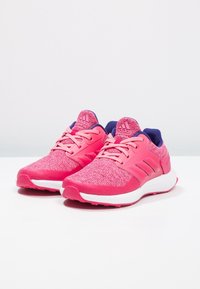 Pár růžových běžeckých bot Adidas s bílou podrážkou a tmavě modrou podšívkou, vystavených na bílé ploše na světle šedém pozadí.