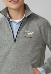 Grijze ritssweater met hoge kraag, voorzien van een gestructureerde stof, voorzakken en geborduurde tekst in zwart en wit.