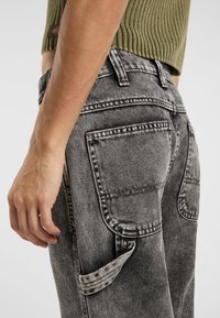 Pantalones de mezclilla gris con un acabado texturizado y desteñido. Presenta dos bolsillos traseros y un bolsillo lateral con un ribete estampado. Primer plano de la cinturilla y la mano.
