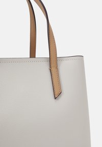 Primo piano di una borsa tote in pelle grigia chiara con una texture, dotata di manici in pelle beige cuciti al bordo superiore, su uno sfondo bianco.