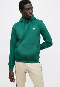 Zelená mikina s kapucňou z bavlny s kanguriou vreckom a sťahovacou šnúrkou na kapucni. Na hrudi má biele logo Adidas; nosená s béžovými nohavicami.