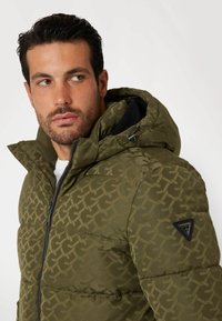 Olive green puffer jacket with a geometric pattern, hood, zipper front, and logo patch on the sleeve. Textured surface and padded design.
