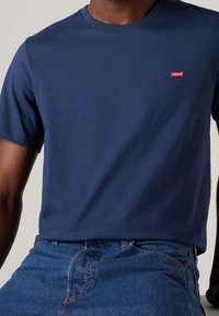 Modrá bavlněná tričko s krátkým rukávem, kulatým výstřihem a malým červeným logem Levi's na hrudi, nošené s modrými džínami.