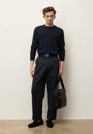 Vyras, vilkintis juodą ilgomis rankovėmis marškinius, tamsias plačias kelnes, juodą diržą, juodus batelius, laikantis juodą pečių krepšį, stovintis prie šviesios sienos.