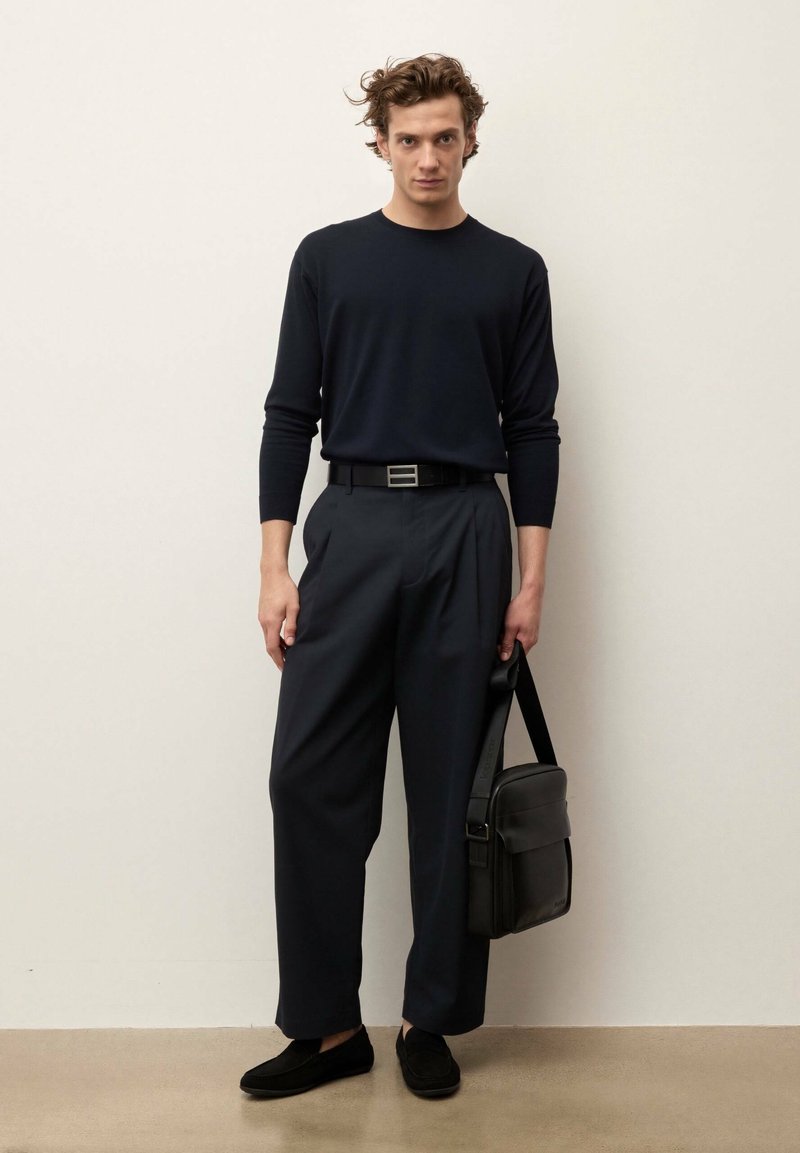 Homme portant une chemise noire à manches longues, un pantalon large foncé, une ceinture noire, des mocassins, tenant un sac à bandoulière noir, debout contre un mur clair.