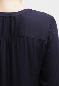 Marinblus med subtil lyster, med v-ringning, rynkad baksida och mjukt draperad tyg för en avslappnad passform.