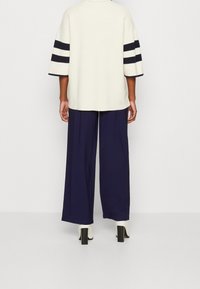 Persona che indossa un maglione color crema con strisce nere sulle maniche, pantaloni ampi blu scuro e stivaletti bianchi con tacco, in piedi di spalle.