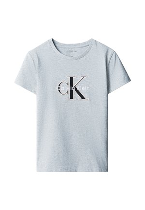 Barn med krøllet hår iført en lysegrå Calvin Klein t-shirt, sorte bukser med vide ben og sorte sko, står mod en ensfarvet baggrund.
