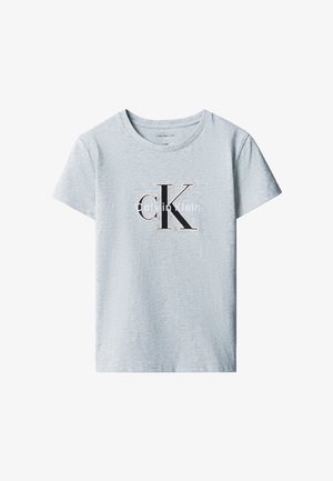 Barn med krøllet hår iført en lysegrå Calvin Klein t-shirt, sorte bukser med vide ben og sorte sko, står mod en ensfarvet baggrund.
