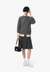 Donna con occhiali da sole neri, felpa grigia sopra una maglietta gialla, gonna plissettata grigia, scarpe da ginnastica bianche e calze, che tiene una borsa nera.