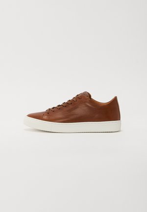 Sneaker de couro castanho com bico arredondado, sola de borracha branca e linhas limpas. Apresenta um fecho com atacadores e uma marcação discreta na lateral.