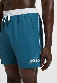 Torso maschile che indossa dei pantaloni da bagno blu teal con cintura e lacci bianchi, tasche laterali e logo "BOSS" sulla gamba sinistra.