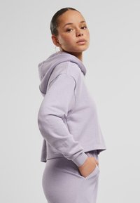 Cappotto corto viola chiaro con una texture morbida, tasche laterali e cappuccio con coulisse, indossato sopra pantaloni coordinati. Orecchini circolari visibili.
