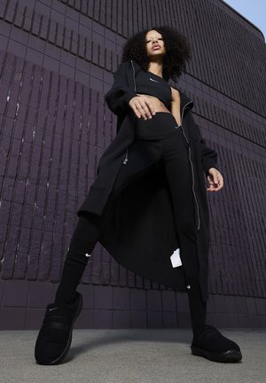 Conjunto atlético negro que incluye una sudadera con cremallera larga, una camiseta corta, leggings de cintura alta, calcetas hasta la rodilla y zapatos negros texturizados, contra un muro púrpura.