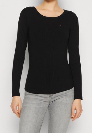 Vrouw draagt een strakke zwarte geribbelde top met lange mouwen met een klein logo op de borst en lichtgrijze spijkerbroek.