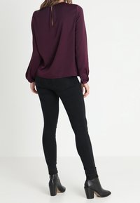 Femme portant un blouse bordeaux à manches longues, un jean skinny noir et des bottines à talons noires, se tenant de dos.