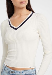 Donna con un top aderente bianco a maniche lunghe e scollatura a V, caratterizzato da una linea nera e bianca, abbinato a jeans blu chiaro.