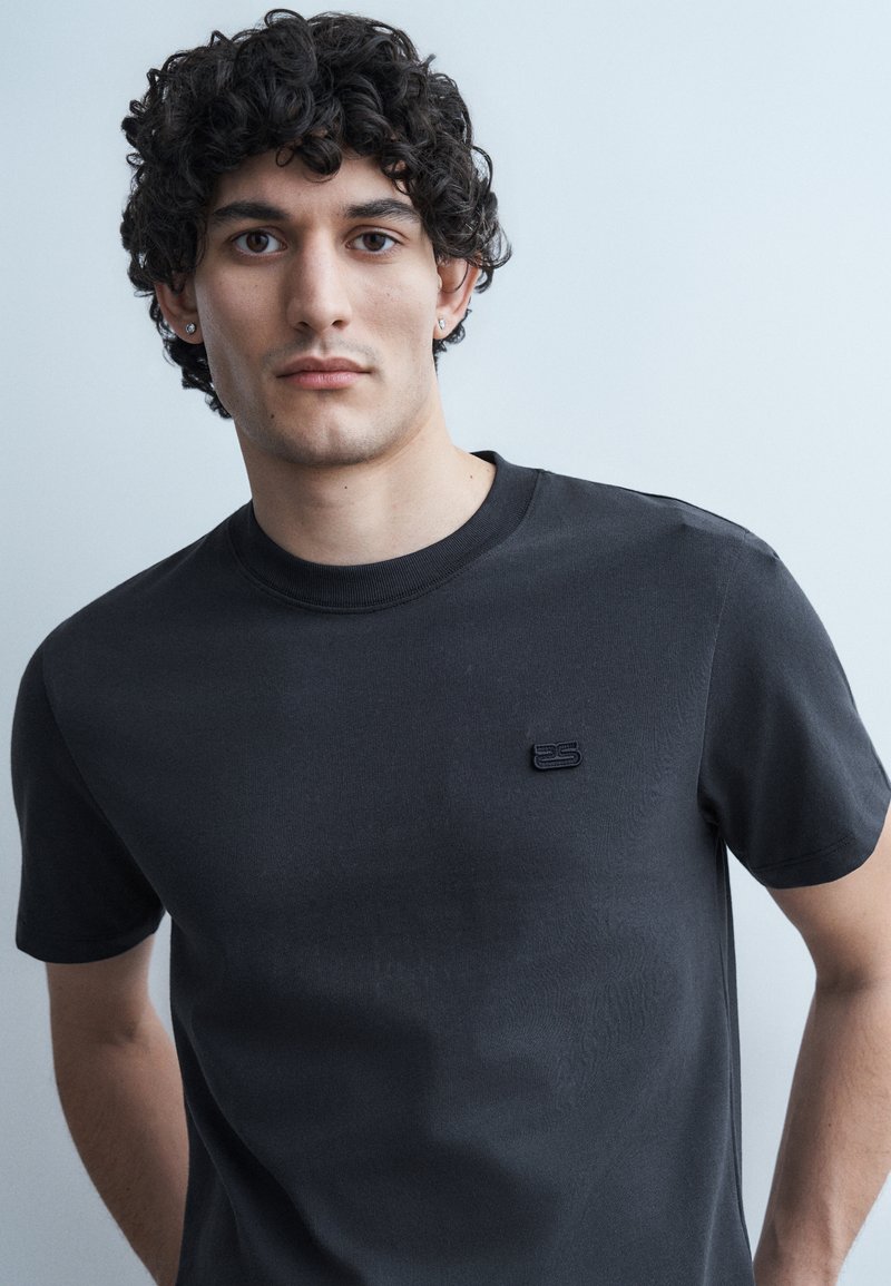 Νεαρός άνδρας με σγουρά σκούρα μαλλιά, που φοράει μαύρο t-shirt με μικρό λογότυπο στο στήθος, στέκεται μπροστά σε ανοιχτό γκρι φόντο.