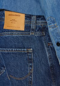 Vaqueros de mezclilla azul oscuro con costuras naranjas, con una etiqueta de cuero que dice "JACK & JONES JEANS INTELLIGENCE." Diseño de bolsillo estándar.