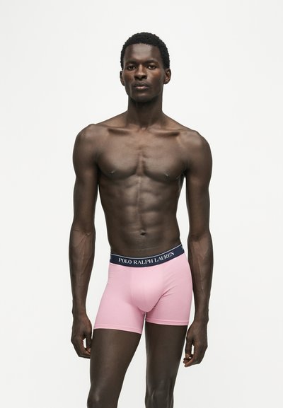 Μυώδης άνδρας στέκεται φορώντας ροζ boxer briefs Polo Ralph Lauren με ναυτικό μπλε μέση, σε απλό λευκό φόντο.