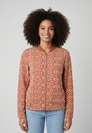 Donna con capelli ricci che sorride, indossa una giacca con cerniera dai motivi geometrici rustici e beige e jeans azzurri, in piedi davanti a uno sfondo semplice.