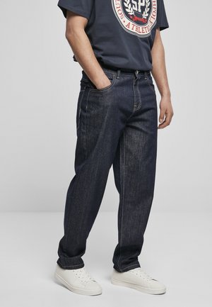 Personne portant un jean droit bleu foncé, un t-shirt graphique bleu marine et des baskets blanches, debout avec une main dans la poche devant un fond uni.