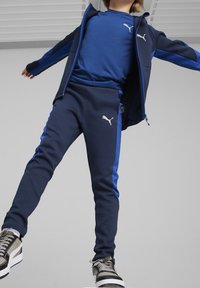 Completo sportivo blu navy con accenti blu, composto da una giacca con zip e pantaloni con vita elastica, completato da sneakers grigie.