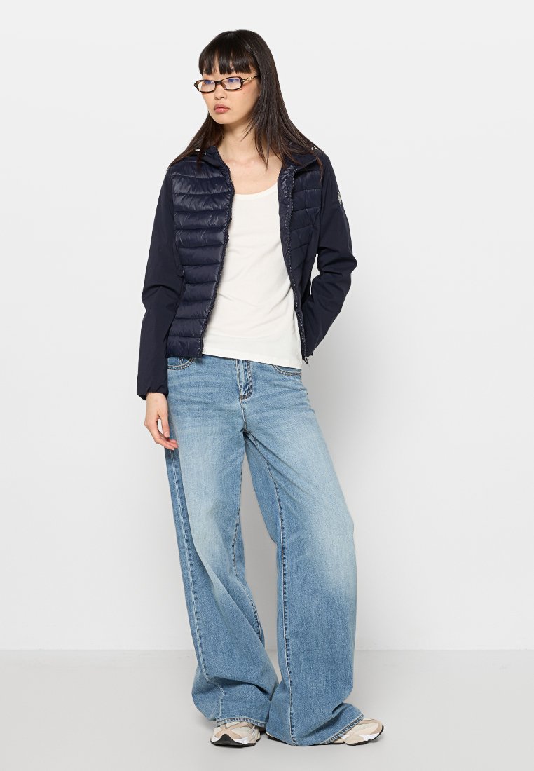 Jonge vrouw met een bril, navyblauwe pufferjack, witte top, lichtblauwe wijde jeans en sneakers, staand tegen een witte muur.