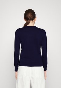 Mörkblå stickad tröja med långa ärmar, ribbade ärmslut och nederkant, med rund halsringning och en mjuk textur. Bärs med vita byxor.