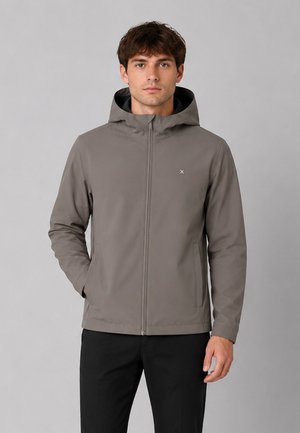 Jeune homme portant une veste à capuche grise avec une fermeture éclair à l'avant et un pantalon noir, debout devant un fond gris clair uni.