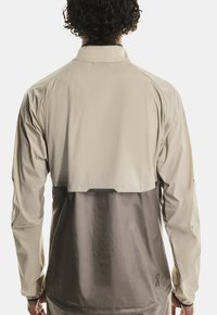 Personne portant une veste légère beige et grise avec un rabat de ventilation sur le dos, vue de derrière.