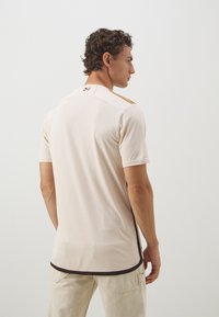 Maglietta sportiva beige con maniche corte, caratterizzata da un collo rotondo e bordi neri a contrasto sull'orlo. Texture sottile, senza motivi visibili.