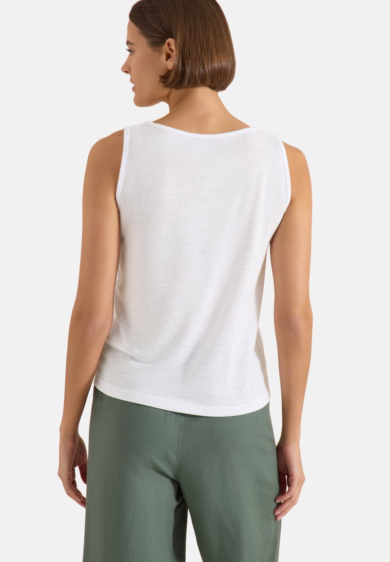 Femme portant un haut blanc sans manches et un pantalon vert, vue de dos sur un fond clair uni.